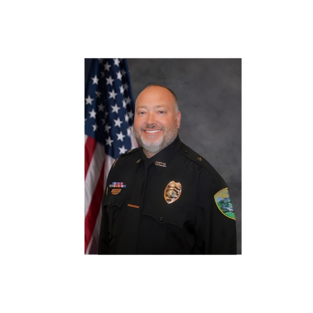 A uniformed police officer with a badge and service ribbons is smiling in front of an American flag.